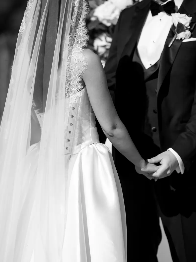 A couple hold hands during the ceremony, the bride's lace-edged veil falling across her shoulder.